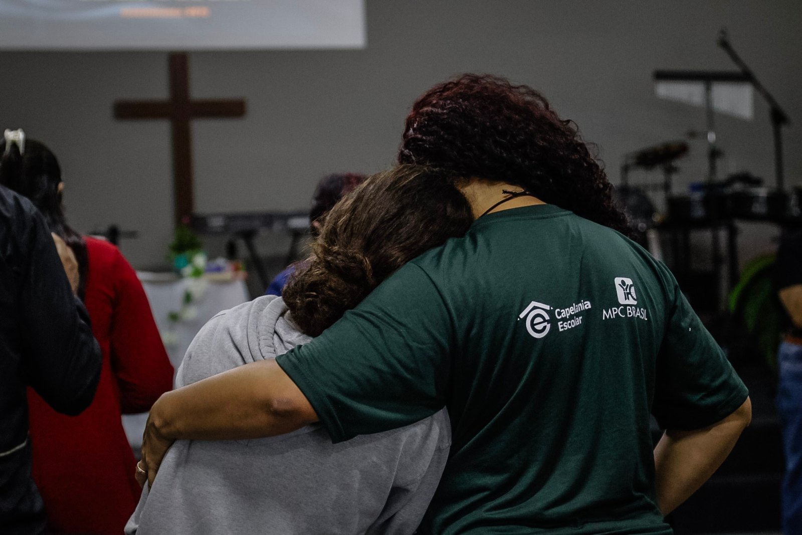 Abraço entre participantes com camiseta da Capelania Escolar MPC Brasil, em frente à cruz